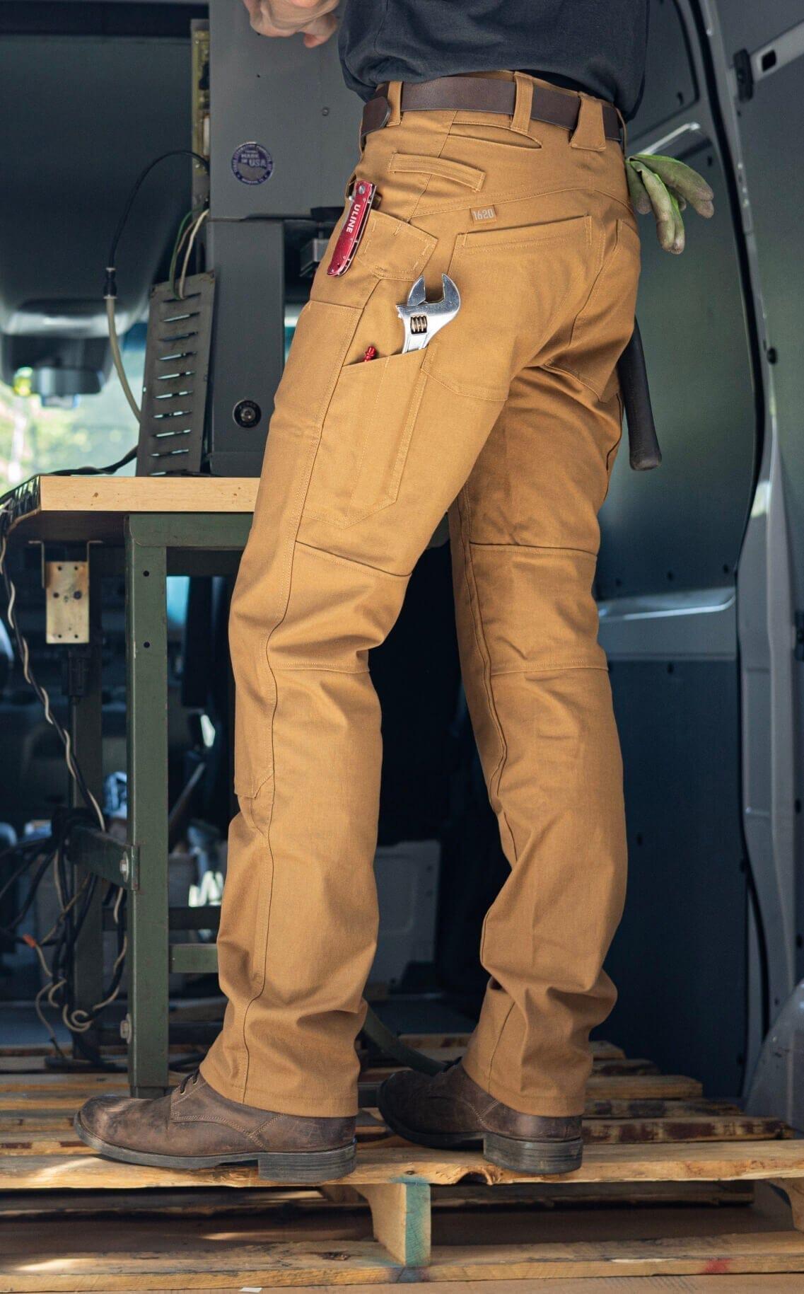 A tradesman standing in a work van wearing tan 1620 USA flexible work pants, showcasing reinforced utility pockets holding a wrench, designed for the demands of electricians and plumbers.